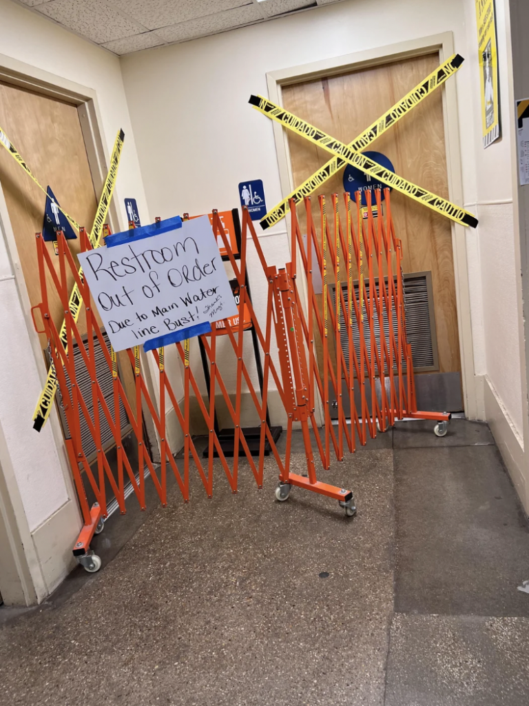 A restroom is blocked off by orange barricades and caution tape. A handwritten sign on the barricade reads "Restroom Out of Order. Due to main water line break!" The area appears to be in a public building, such as a school or office.