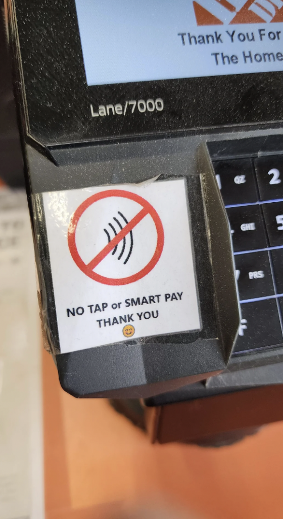 Close-up of a card payment terminal with a sign showing a contactless payment symbol crossed out. The text on the sign reads, "NO TAP or SMART PAY THANK YOU" followed by a smiley face emoji. The terminal model is "Lane/7000.