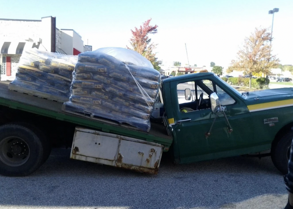 A green pickup truck with a tilted flatbed is loaded with multiple bags of concrete mix. The truck appears to be struggling under the weight, causing the flatbed to tilt significantly. In the background, there are trees, a building, and a clear sky.