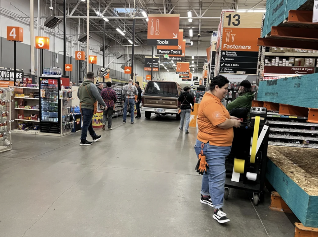 Customers shop in a hardware store with tall shelves filled with various tools and supplies. An employee in an orange apron assists a customer near aisles marked 13 and 14. In the background, more customers browse and a vintage truck is partially visible.