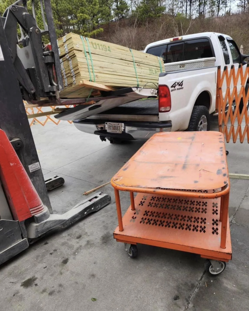 A forklift is loading a bundle of wooden planks onto the bed of a white pickup truck. An orange mobile cart is positioned nearby. Safety barriers are partially visible, along with a few trees and a concrete surface in the background.