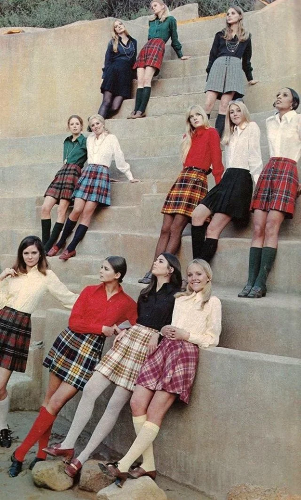 A group of women are standing and sitting on large concrete steps. They are dressed in various colored plaid skirts, knee-high socks, and solid-colored tops. Their attire and hairstyles suggest a vintage or retro fashion style. The setting appears to be an outdoor area.