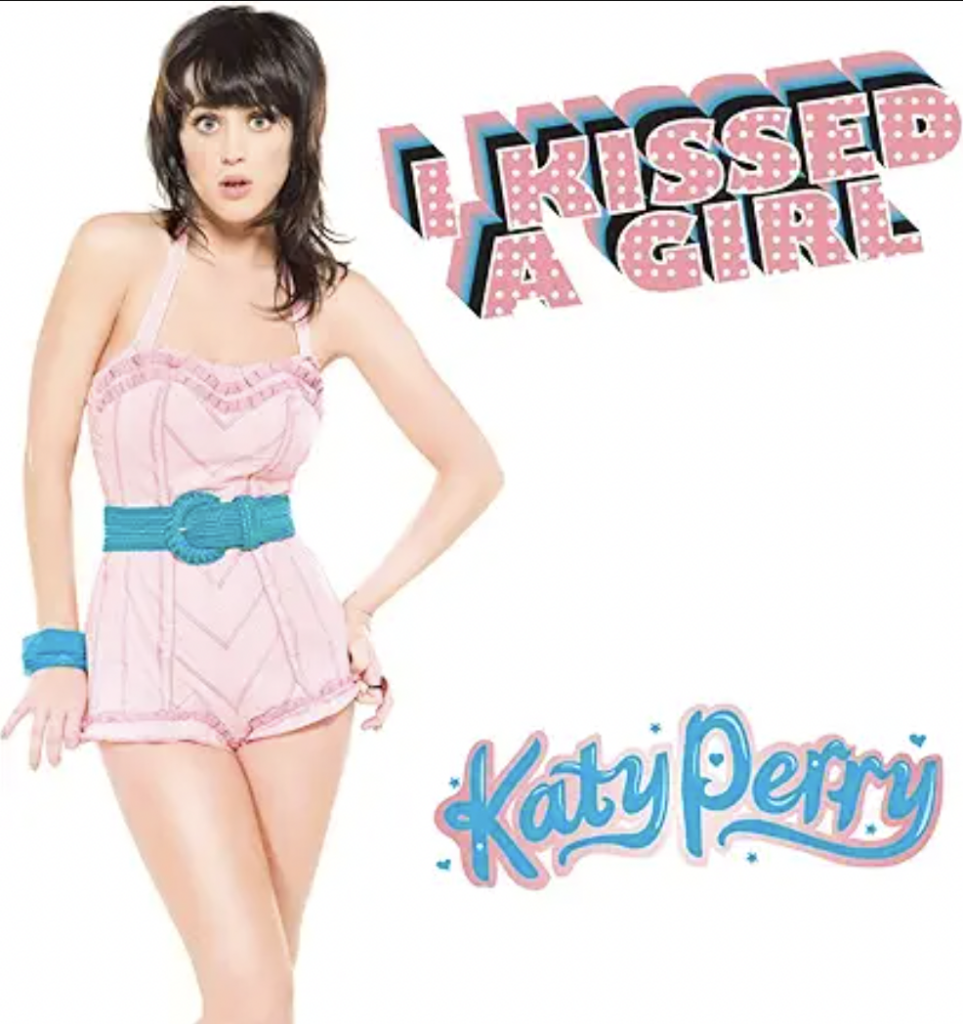 A woman with dark hair, styled in a bob cut, is wearing a pink retro-style romper with blue accessories. Above her, colorful 3D text reads "I Kissed a Girl" and below her, in a similar style, "Katy Perry" is written.