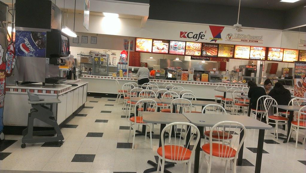 A cafeteria with numerous empty tables and white chairs with orange seats. A few customers are seated at the far right. The counter in the background features a variety of food and drink options, with brightly lit menu boards overhead.