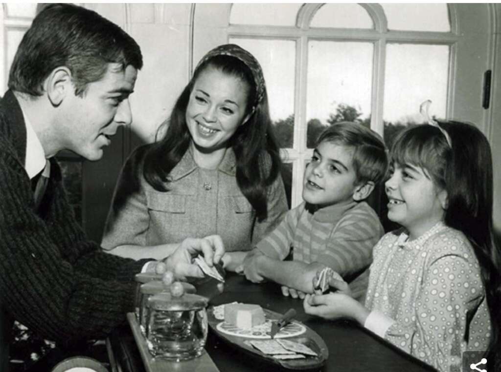 A black and white photo shows a man, a woman, a boy, and a girl sitting together at a table. The man and woman are smiling at the children. On the table are some plates and a glass. The family seems happy and engaged in conversation.