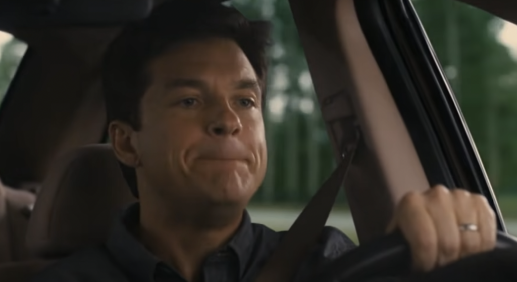 A man with short dark hair is driving a car. He is wearing a seatbelt and looks concerned or deep in thought. The blurred background outside the car window suggests movement.