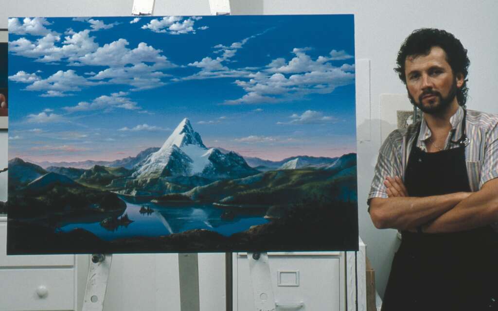 A man stands beside an easel holding a completed painting. The painting depicts a serene landscape with a towering snow-capped mountain reflected in a tranquil lake, surrounded by rolling green hills under a sky dotted with fluffy clouds. The man has curly hair and wears a striped shirt and apron, with arms crossed.