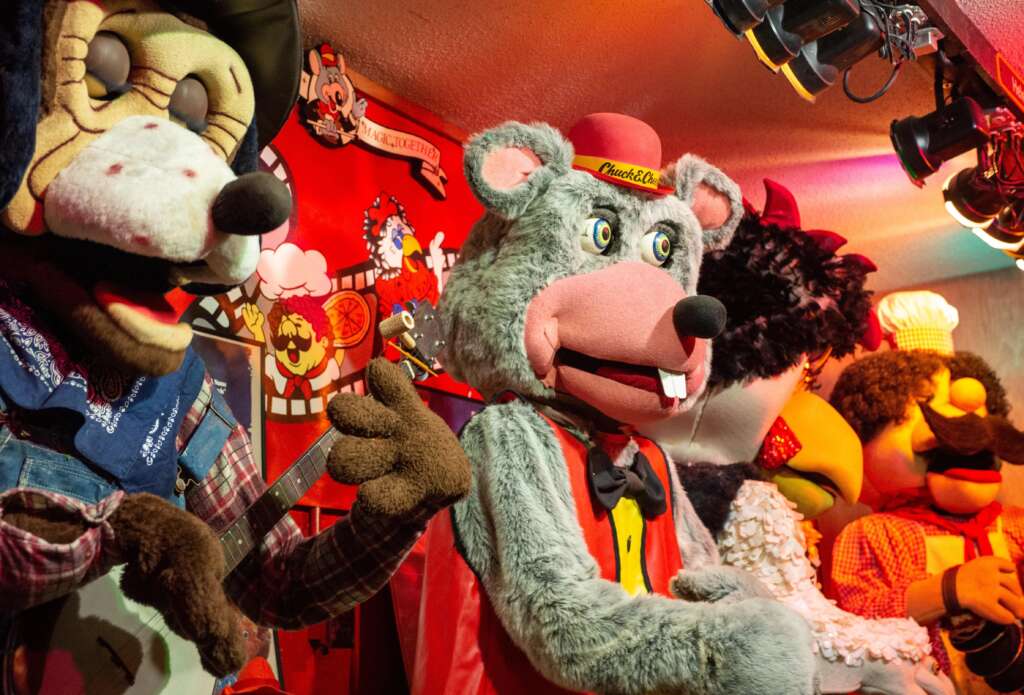 A group of colorful animatronic characters performs on a vibrant stage. One character, a gray mouse in a red vest and yellow shirt, is prominent. The background is adorned with various bright decorations and lights, creating a lively and playful atmosphere.
