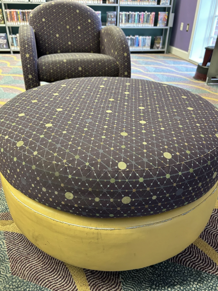 A cushioned round ottoman with a patterned fabric and a yellow base is placed in the foreground. Behind it, there is a matching armchair with similar patterned upholstery. In the background, bookshelves filled with books are visible.