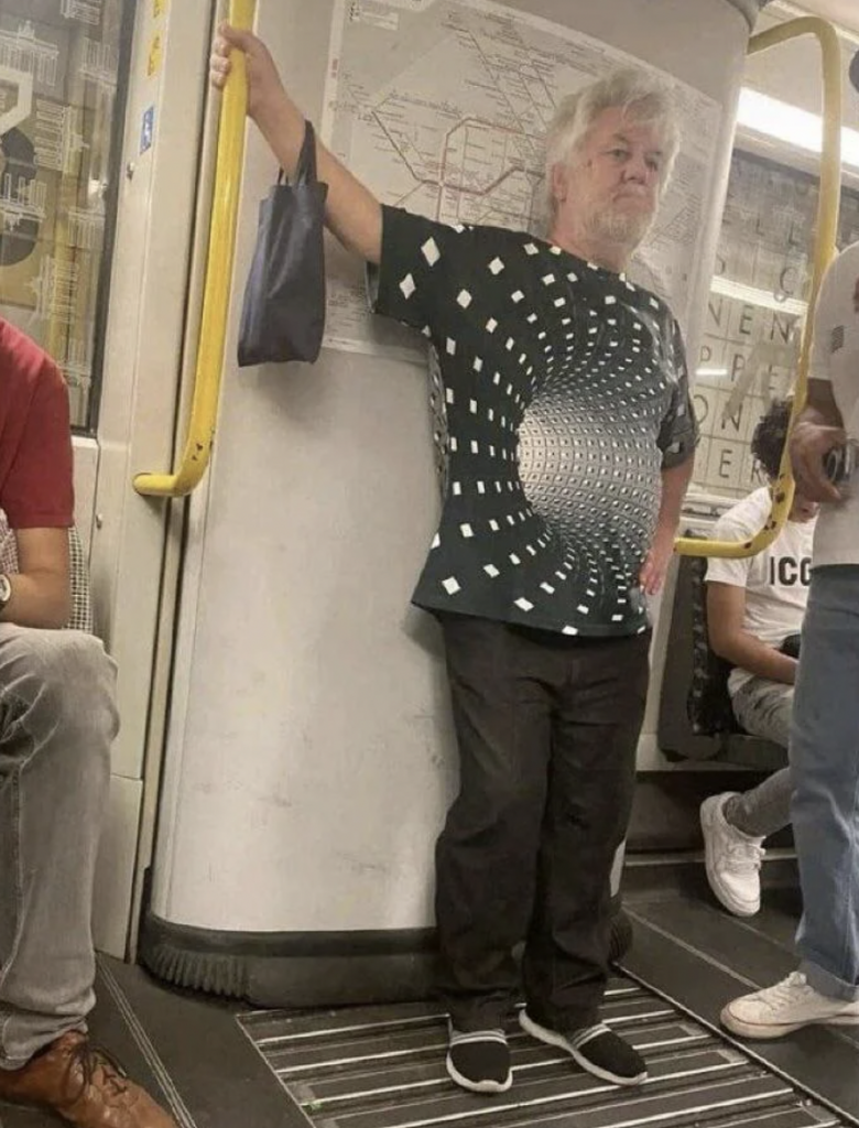 A man stands on a subway train, holding onto a yellow pole. He is wearing a shirt with an optical illusion pattern. Another passenger in a red shirt is seated nearby, and part of an underground map and additional passengers are visible in the background.