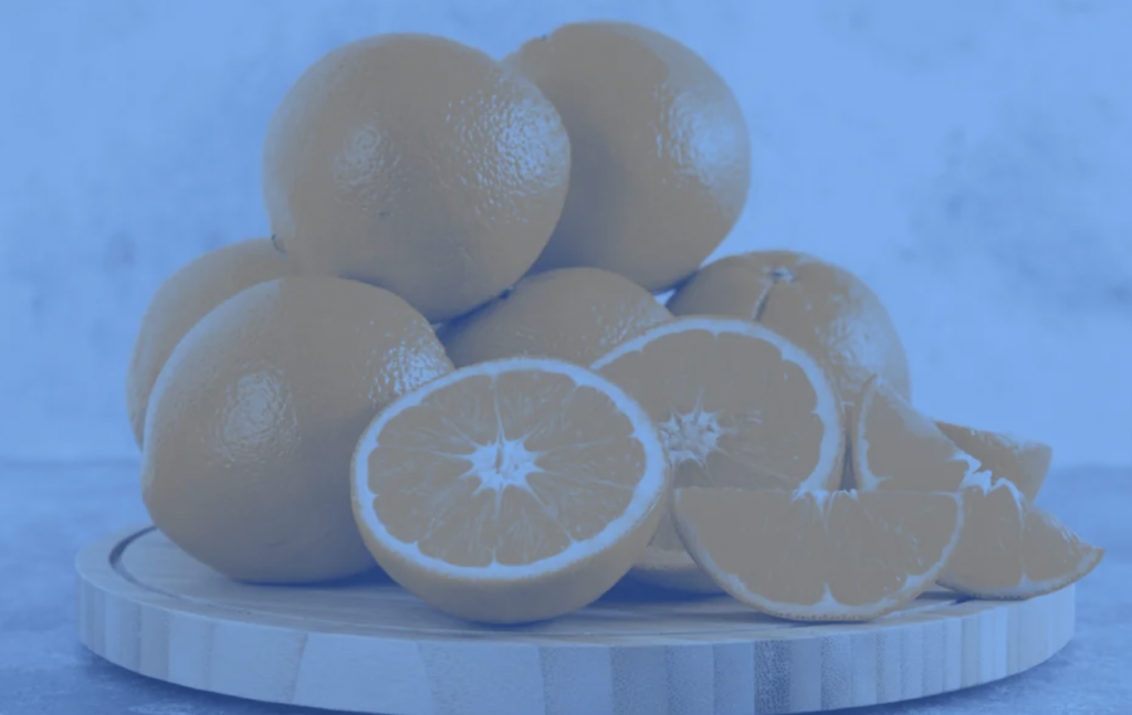 A stack of whole oranges with several cut in half and into wedges on a round wooden board. The entire image has a blue tint. The background is a textured pale blue surface.