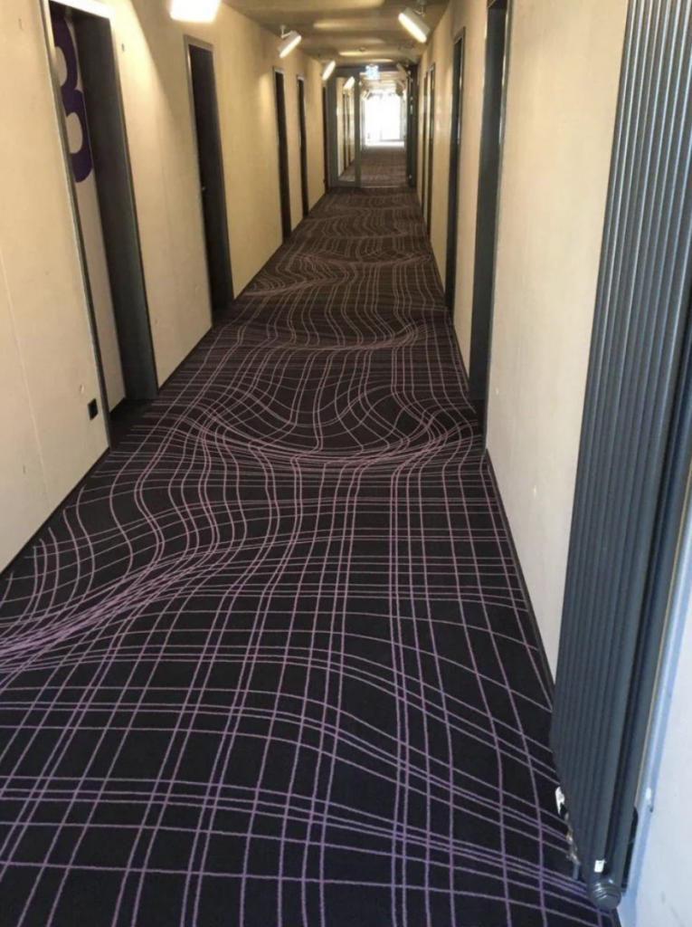 A hallway with a carpet featuring an optical illusion: it appears to have wavy distortions in the pattern, creating a visual effect that makes the floor look uneven. Doors line both sides of the hallway, and overhead lights illuminate the corridor.