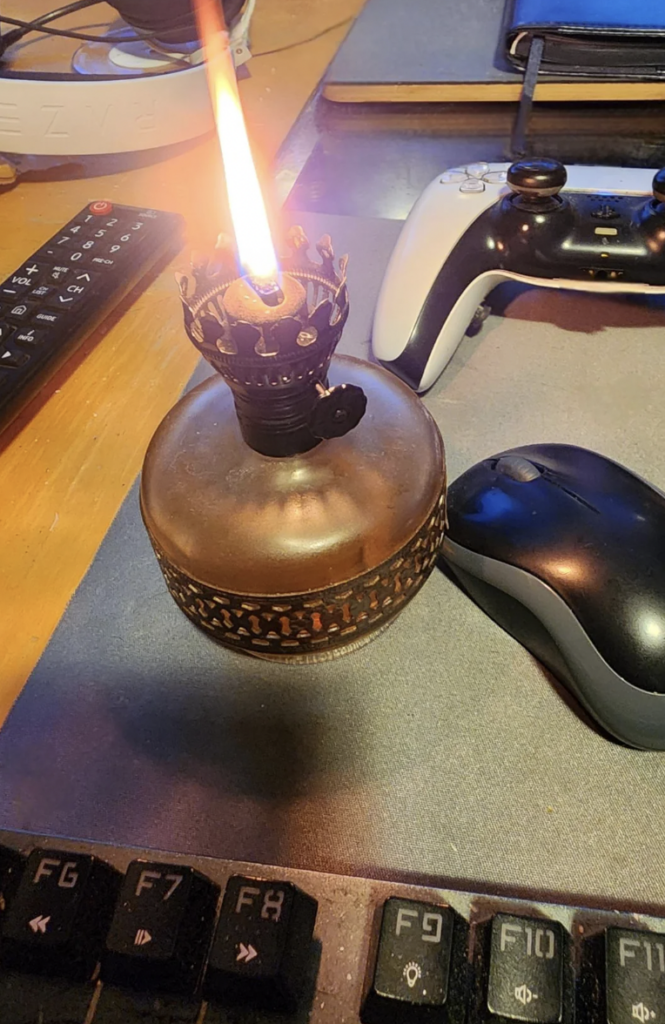 A lit oil lamp with a decorative metal crown sits on a desk alongside a remote control, two game controllers (one white, one black), a computer mouse on a mouse pad, and a keyboard. The scene is warmly lit, highlighting the lamp's flame.