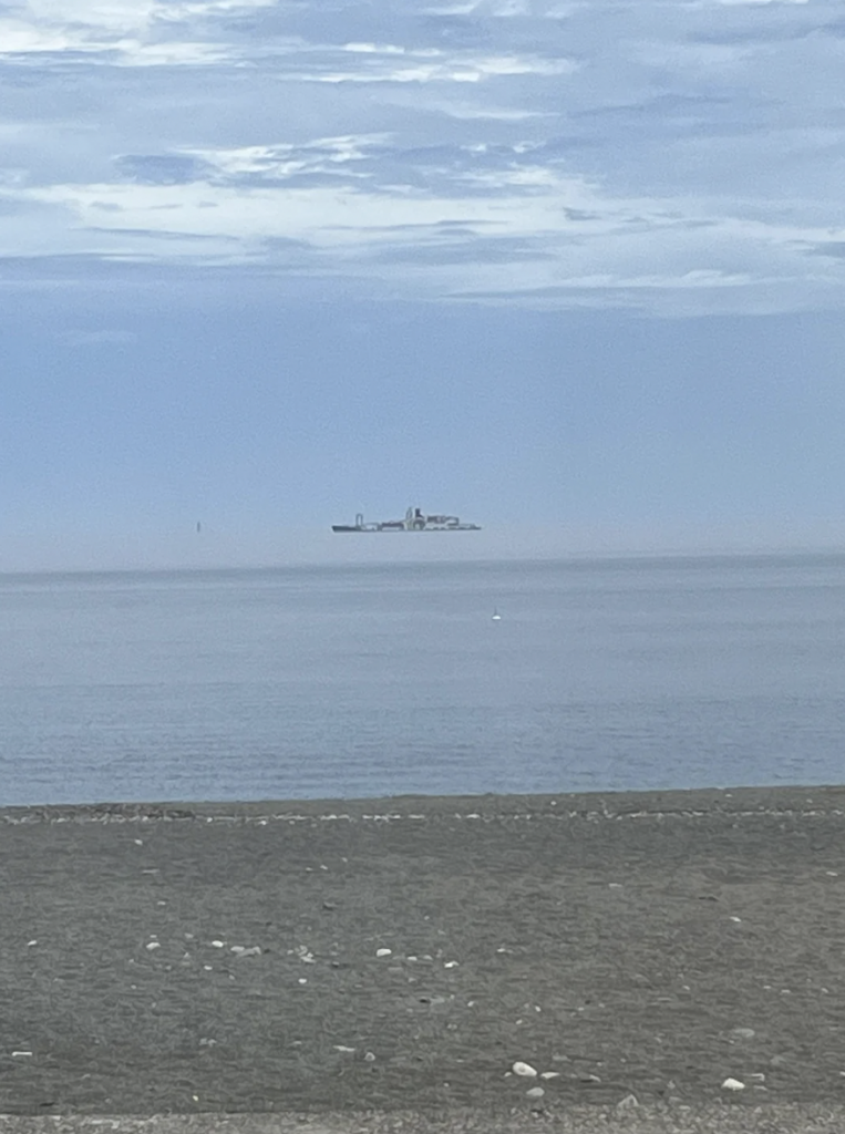 A large ship appears to be floating above the horizon against a blue sky. The optical illusion creates a surreal effect. The ship is located in the distance over calm ocean waters, with a pebbled beach in the foreground.