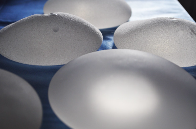 Close-up image of several transparent silicone breast implants placed on a blue cloth surface. The implants vary in shape and size, with a textured surface that scatters light, giving a frosted appearance.