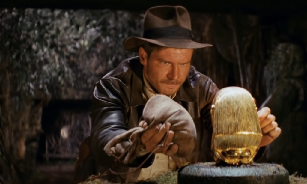 A man wearing a brown leather jacket and fedora cautiously swaps a sandbag for a golden idol in an ancient, dimly-lit temple. The idol is placed on a stone pedestal, and he appears focused and tense during the exchange.