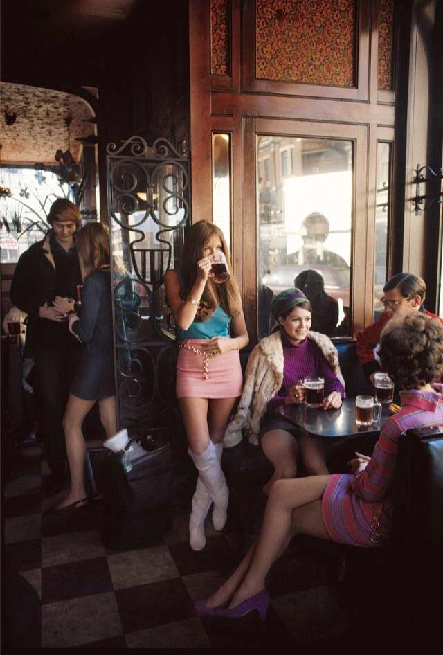 A stylish woman in a pink skirt and knee-high boots drinks beer by a bar's window. Nearby, two women and a man enjoy drinks at a small table. Another woman stands and chats with a man by a doorway. The scene captures a vintage, lively social atmosphere.