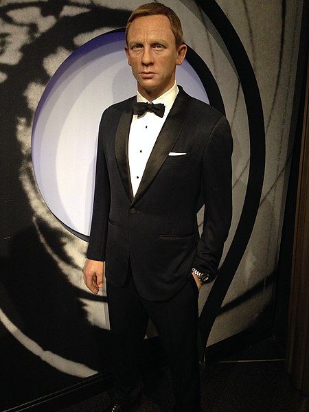 A wax figure of a man in a black tuxedo with a bow tie stands against a backdrop resembling a gun barrel design from the James Bond film series. The figure has short, neatly combed hair and a serious expression.