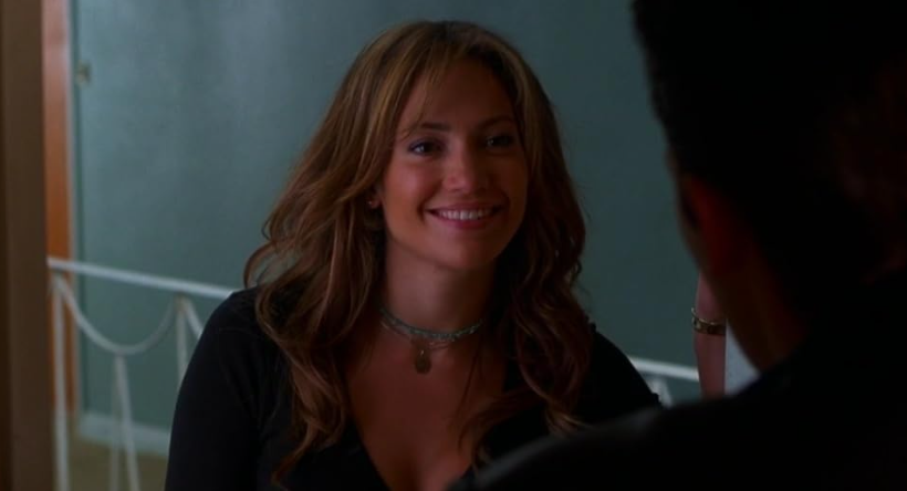 A person with long, wavy, brown hair is smiling and looking to the right. They are wearing a black top and a necklace with a pendant. The background features a neutral-colored wall and part of a railing.