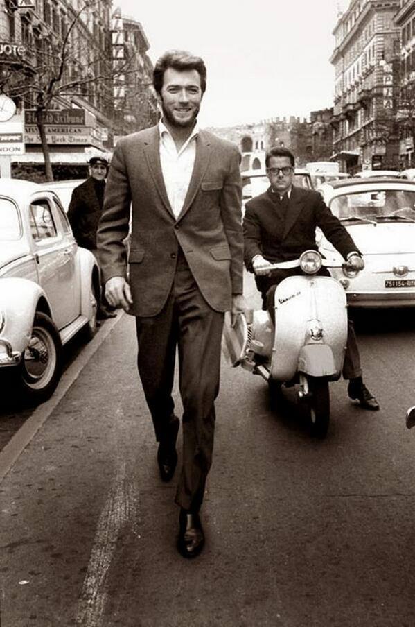 A black and white photo of a man in a suit and open-collared shirt smiling as he walks down a busy street. Behind him, a man rides a Vespa scooter, and several cars, including a vintage Volkswagen Beetle, are visible. The street is lined with tall buildings.