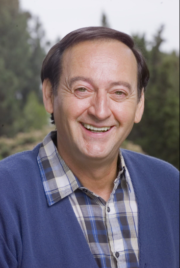 A man with short dark hair is smiling widely at the camera. He is wearing a blue plaid shirt underneath a blue cardigan. The background is slightly out of focus, with green trees and plants visible, suggesting an outdoor setting.