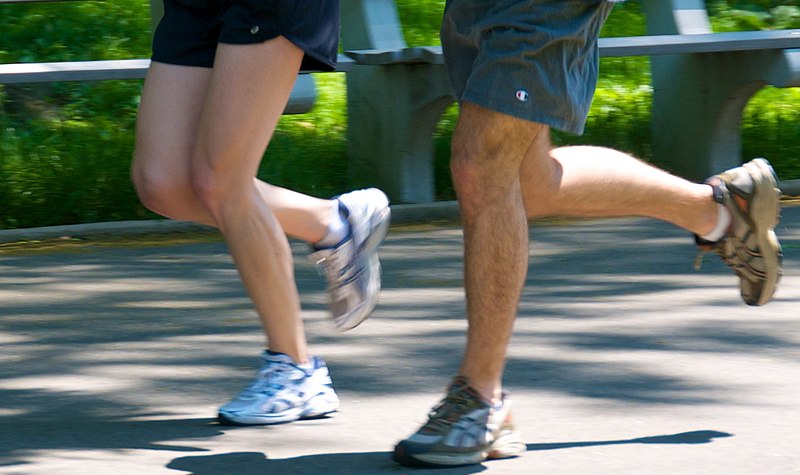 Close-up of two people jogging side by side on a sunny day. Their faces are not visible, and only their legs and feet in motion are shown. Both are wearing athletic shoes, shorts, and the background features blurry greenery and a bench.