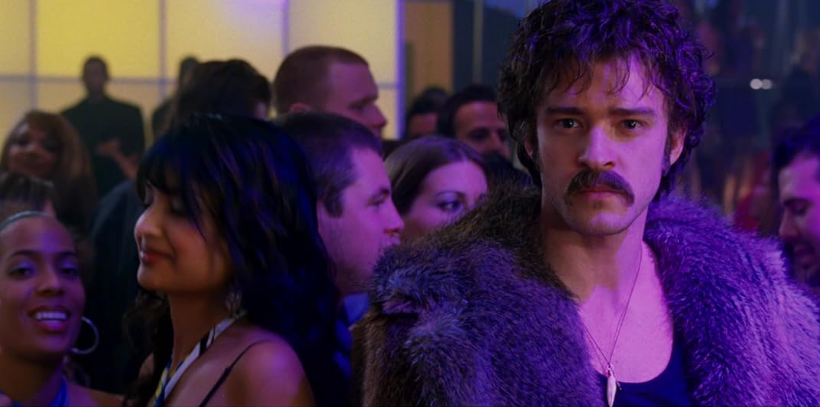 A man with curly hair, a mustache, and a fur coat stands in a crowded, dimly lit room with colorful lighting. People around him are smiling and talking, creating a lively atmosphere. The man looks directly at the camera with a serious expression.