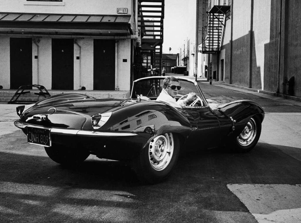 Black and white photo of a person driving a vintage convertible sports car, with text "JNH 809" on the license plate. The person is wearing sunglasses and looking back over their shoulder. Buildings and an external staircase are visible in the background.