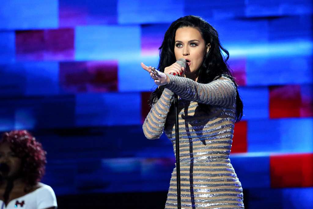 A woman with long black hair, wearing a shimmering silver and gold striped dress, is singing into a microphone on stage. She extends her arm towards the audience. The background is a colorful, abstract pattern with shades of blue, purple, and red.