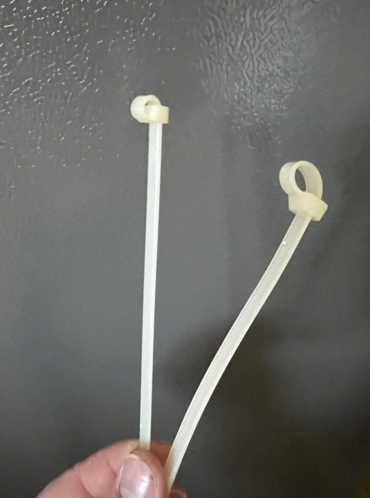 A hand holding two white cable ties in front of a textured gray background. The cable ties are looped but not fully tightened.