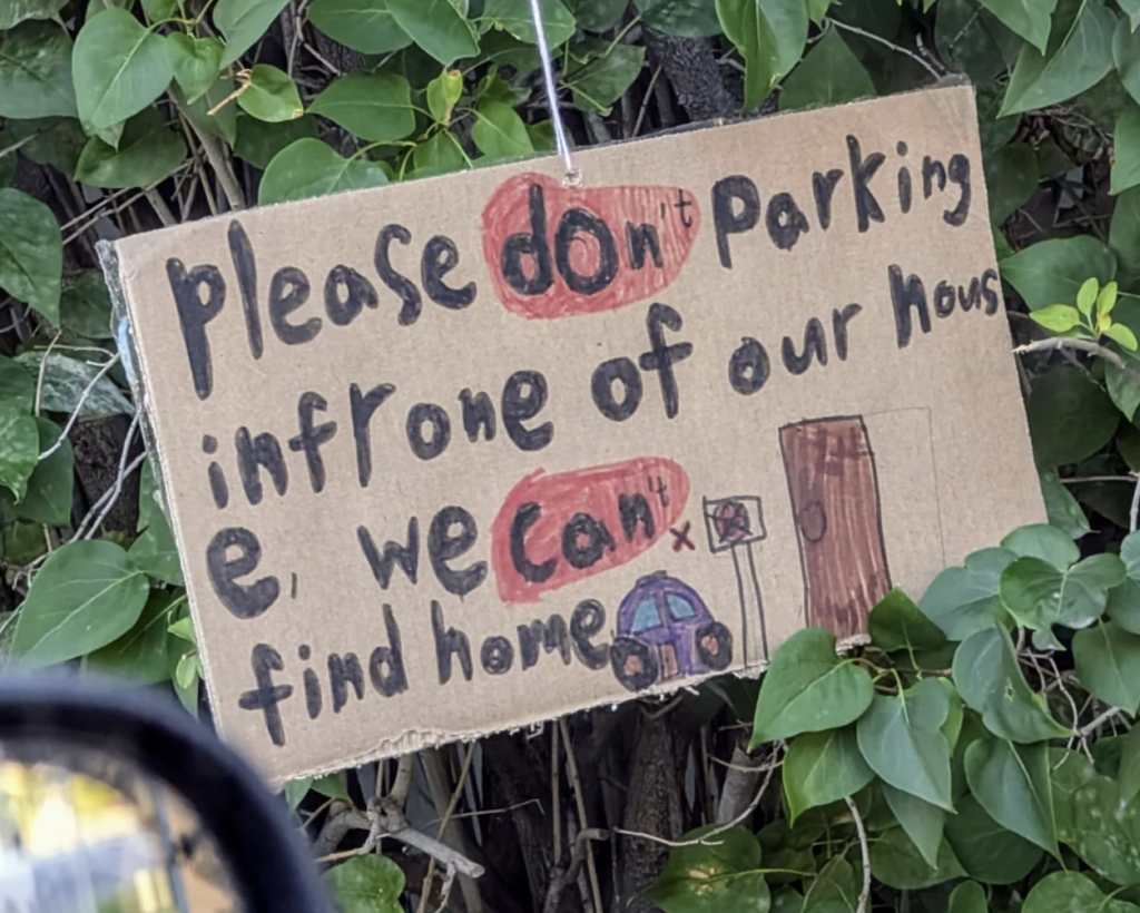 A handwritten sign on cardboard is hung on a leafy background. The message reads, "Please don't parking in front of our house, we can't find home," with drawn images of a house, a car, and red circles around the words "don't" and "can't.