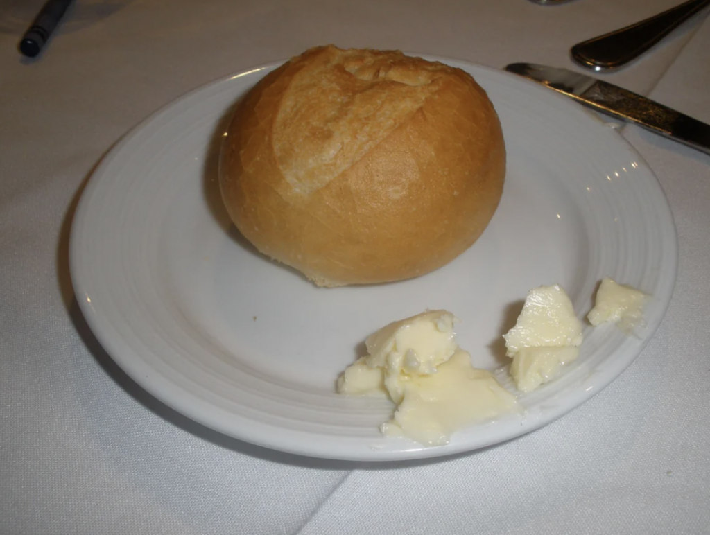 A round bread roll placed on a white plate next to a couple of small butter chunks. The plate is set on a white tablecloth with a knife beside it.