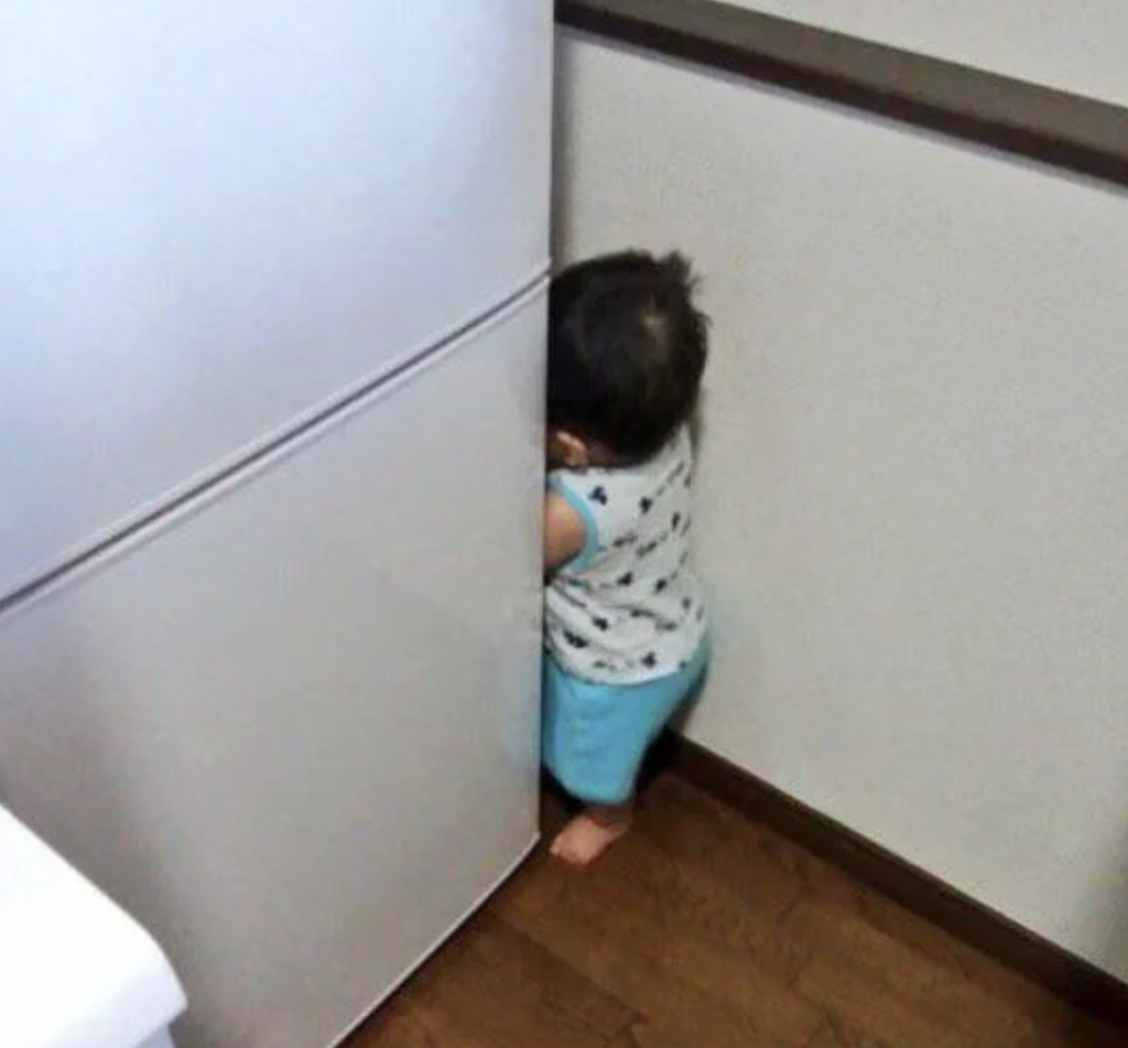 A small child in a sleeveless shirt and blue pants stands between a refrigerator and a wall, facing the wall, creating a humorous and endearing scene as if playing or hiding in the tight space.