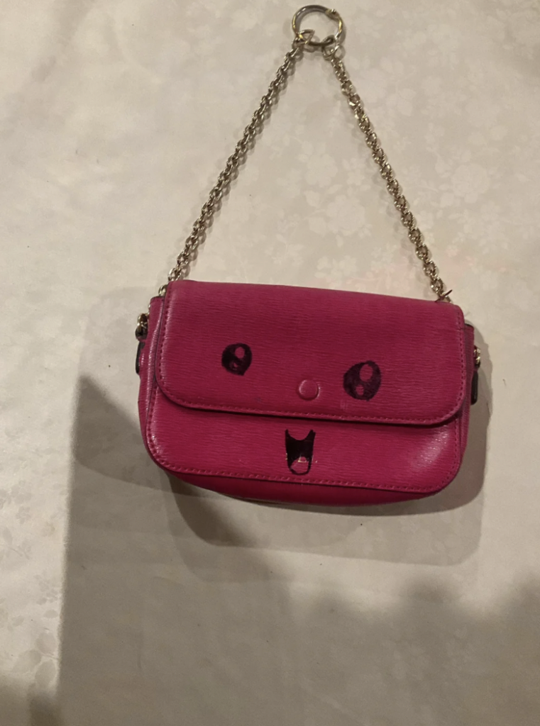 A small bright pink handbag with a chain strap. The front flap has a cute face design with large, wide eyes and an open, smiling mouth. The background is a light, textured surface.