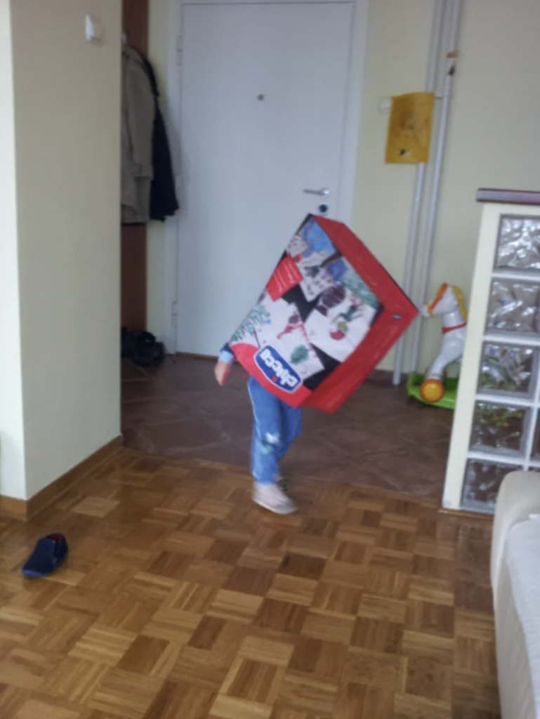 A child with a large rectangular cardboard box over their upper body and head, standing in a hallway at home. The box features various images and text, and the child is barefoot with one shoe visible on the floor. Toys and a coat are in the background.