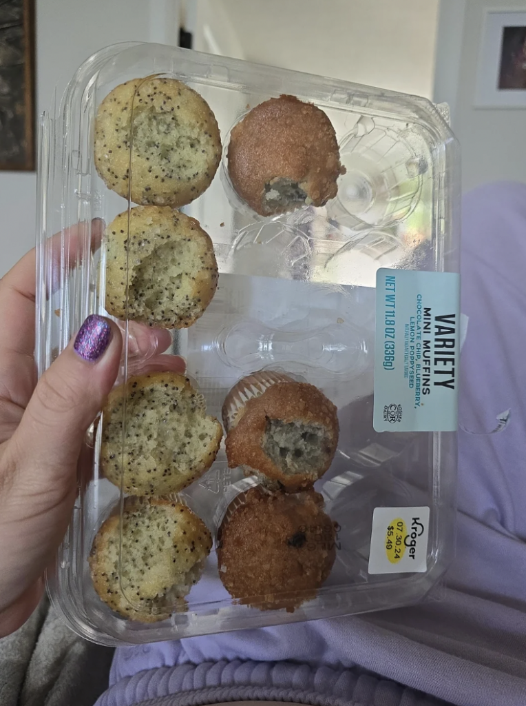 A person is holding a plastic container of assorted mini muffins with several already eaten or missing. The muffins appear to have different flavors, such as poppy seed and chocolate chip. The person's nails are painted a sparkling purple, and they are wearing light purple pants.