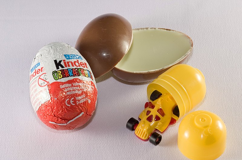 An unopened Kinder Surprise egg lies next to an unwrapped, split-open one, revealing chocolate halves and a yellow plastic capsule toy. The toy, resembling a small, yellow vehicle with red accents, is partially visible next to the capsule.