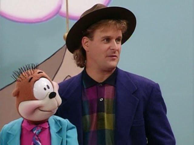 A man with curly hair, wearing a brown hat, blue blazer, and a colorful plaid shirt, stands next to a puppet. The puppet resembles an animal, has spiky hair, and is dressed in a blue jacket and pink tie. The background features a simple, pastel-colored drawing.