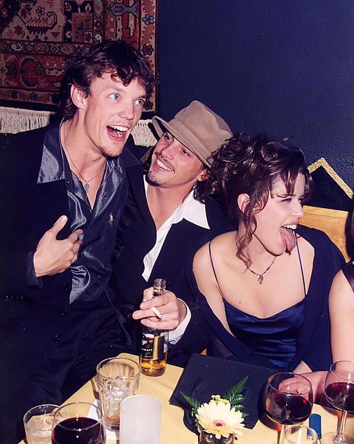 Three people are sitting at a table in a restaurant or bar. One person on the left is energetically talking with their mouth open, another in the middle is smiling warmly and wearing a hat, and the third person on the right is sticking their tongue out playfully.
