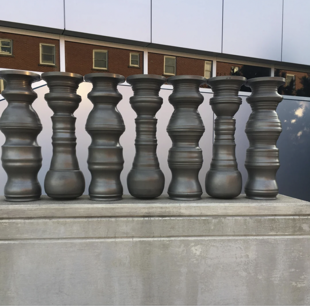 A row of seven large, metallic, differently designed spindle-shaped sculptures is mounted on a concrete base. Behind them, a reflective wall partially shows the facade of a building with several windows.