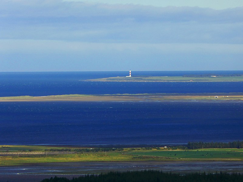 A picturesque coastal scene featuring a serene expanse of blue ocean meeting a clear sky. In the distance, a lighthouse stands on a small, low-lying landmass extending into the sea. The foreground displays a contrasting patchwork of green fields and a dark blue water body.