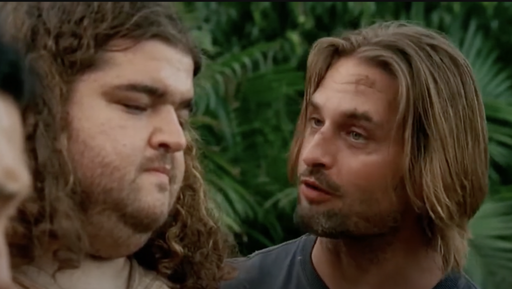 Two men are in a close-up scene set against a backdrop of lush green foliage. The man on the left has long curly hair and a beard, and is looking down with a serious expression. The man on the right has dirty blonde hair and appears to be talking.