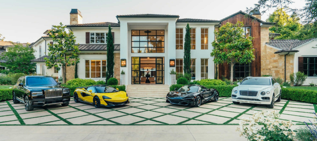 A luxurious mansion with a modern design featuring large windows and a well-manicured lawn. In the driveway are four luxury cars, including a black Rolls Royce, a yellow sports car, a black sports car, and a white Bentley. Trees and plants surround the house.