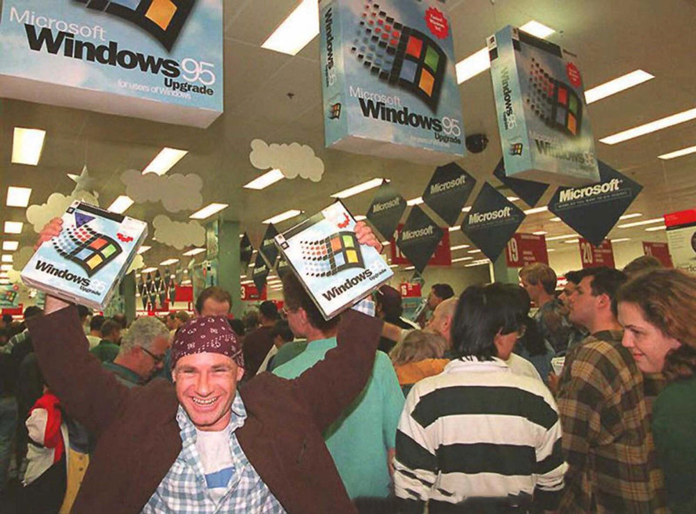 A jubilant man displays two boxed copies of Microsoft Windows 95 in a crowded store. Shoppers around him look engaged in their own purchases. The store is decorated with large Windows 95 signs hanging from the ceiling.