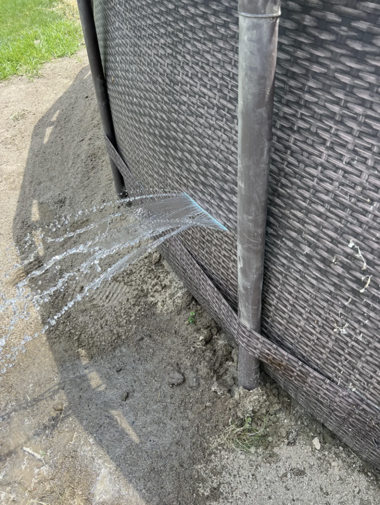 Close-up of an above-ground pool made of wicker-like material with a significant water leak. Streams of water are seen gushing from a crack near the base of the pool, splashing onto the ground below. The surrounding area appears to be wet and muddy.