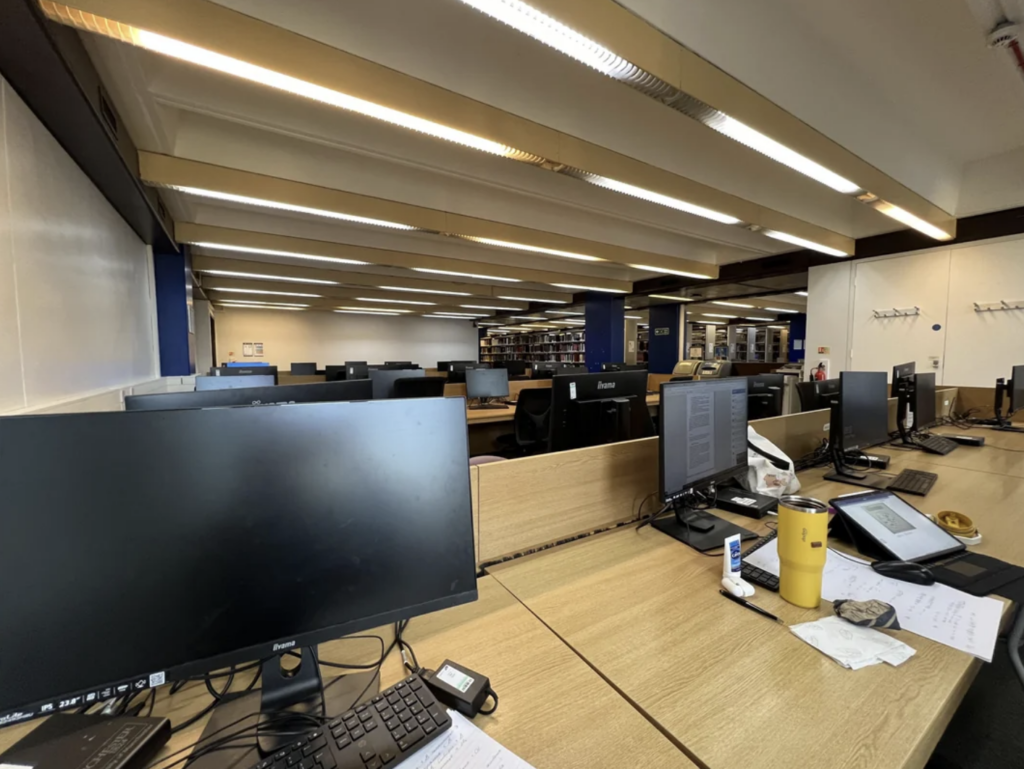 A large office space is filled with rows of empty desks, each equipped with desktop computers, keyboards, and monitors. The desks are separated by dividers, and some personal items like cups and papers are visible. Shelves with books are in the background.