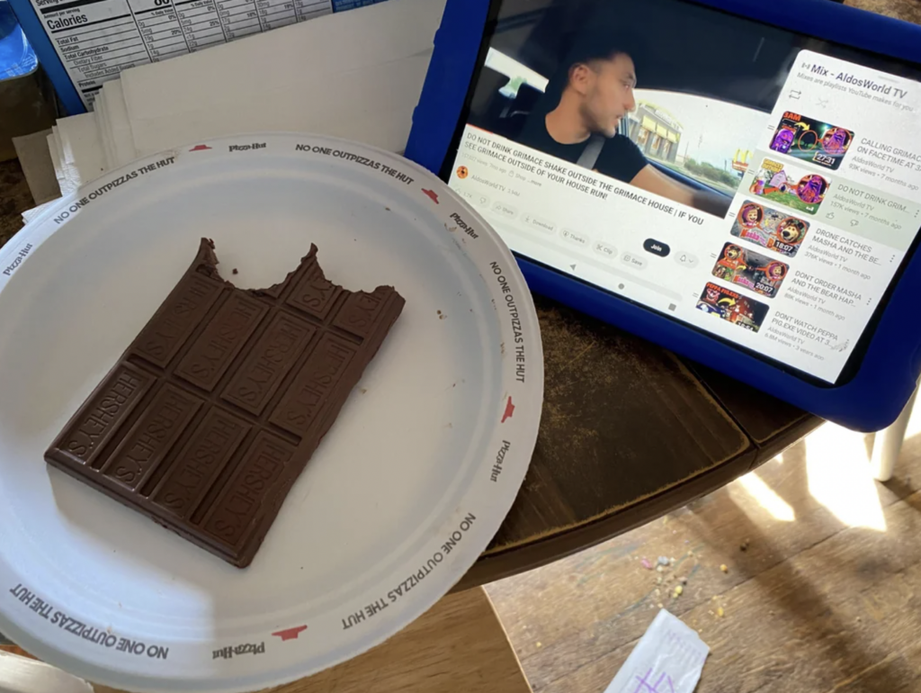 A bitten chocolate bar on a white plate sits on a table beside a blue-framed tablet playing a car vlog on YouTube. There's paper with writing in the background, and sunlight is streaming onto the table. The plate has the text "NO ONE OUTPIZZAS THE HUT.