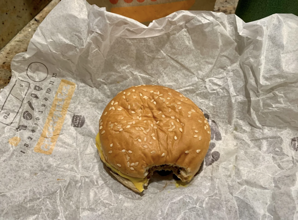 A partially eaten cheeseburger with a sesame seed bun sits on white, crumpled wrapping paper with black and yellow text. The burger reveals layers of meat patty and melted cheese. The background includes fragments of packaging.