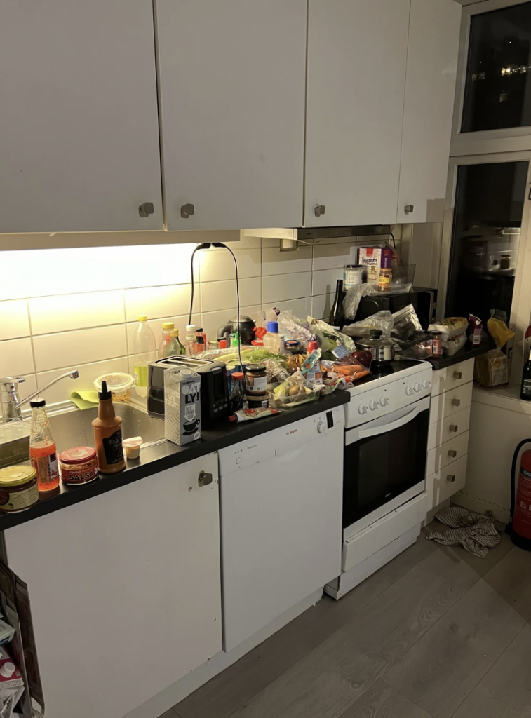 A cluttered kitchen countertop filled with various food items, containers, bottles, and bags. The white cabinets above and below are partially open. The kitchen includes a dishwasher and a stove with an oven. The lighting is dim, except for under-cabinet lighting.