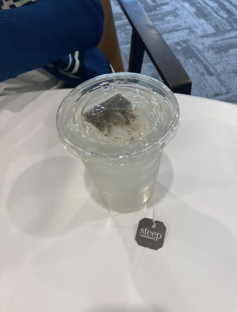 A cold brew tea cup with a teabag hanging inside on a white table. The tag attached to the teabag reads "steep." There is a blue piece of clothing partially visible at the top left corner and a chair's armrest on the right side.